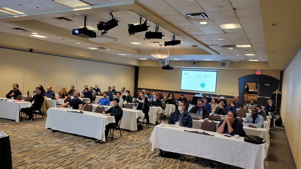 &quot;Five rows of tables with white table clothes with people attentively looking forward at the speaker, who is not in frame, with a PowerPoint slide on the back wall describing solution architecture at the University of Manitoba